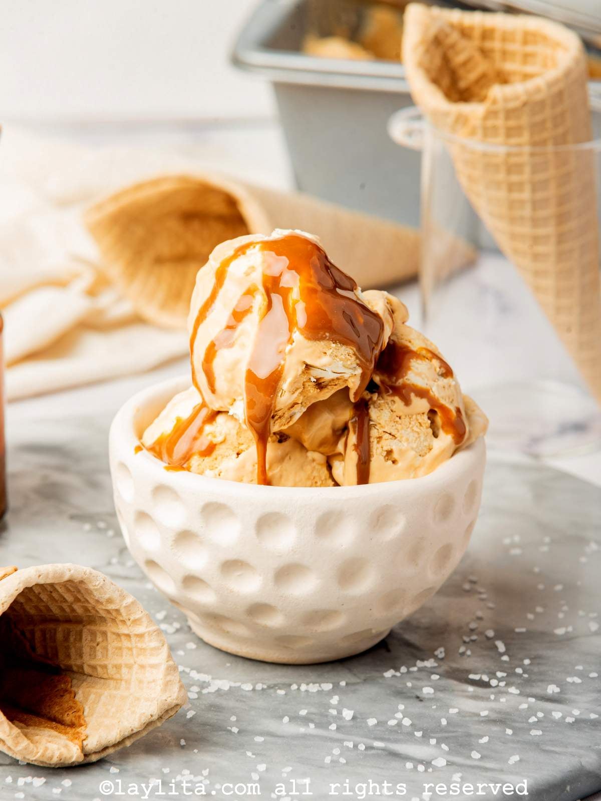 White bowl filled with caramel ice cream with some cones in the background.