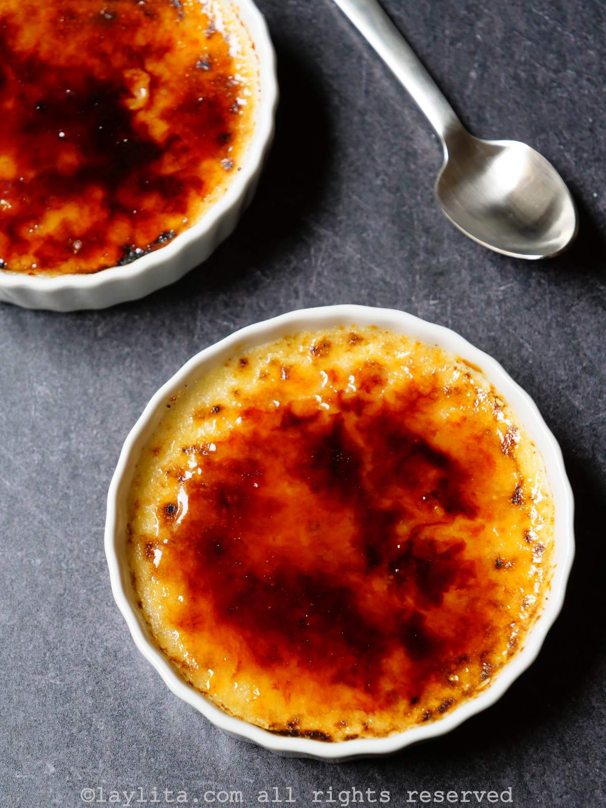 Overhead shot of two ramekins of creme brulee with a caramelized topping, and a spoon on the side