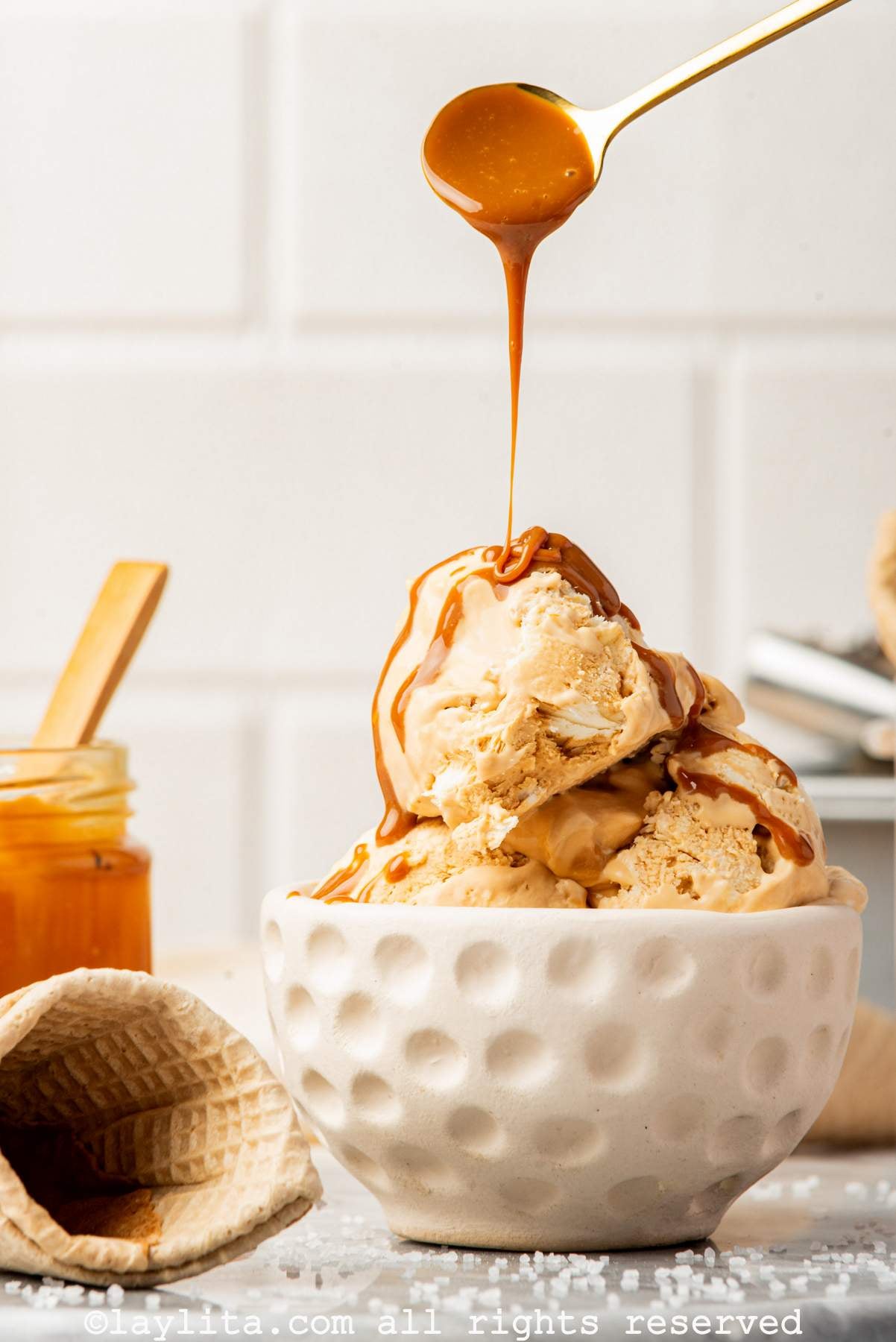 Scoops of salted caramel ice cream in a white bowl with a spoon drizzling more caramel on top.