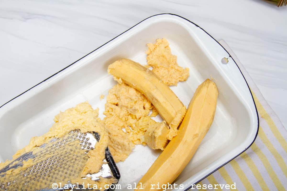 Peeled green plantains being grated into a baking dish.