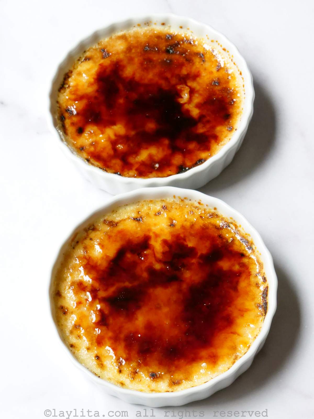 Overhead shot of two ramekins of creme brulee with a caramelized topping on a white background.