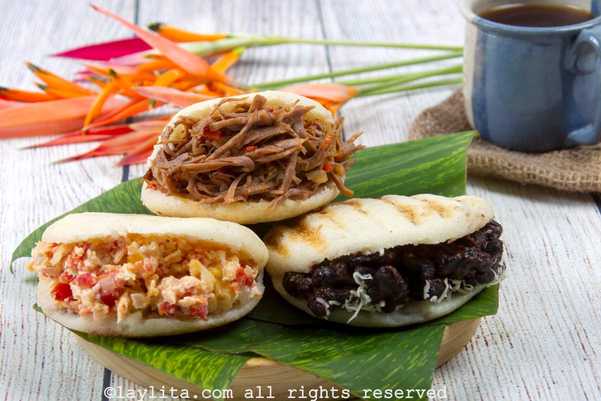 Three stuffed arepas, all stuffed with different fillings, placed on a leaf with a cup of coffee in the background.