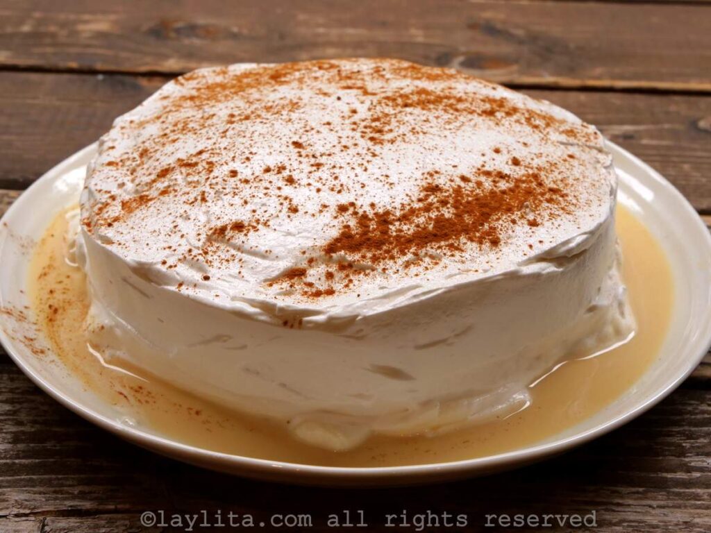 A round tres leches cake on a white plate with frosting and milk mixture on the sides