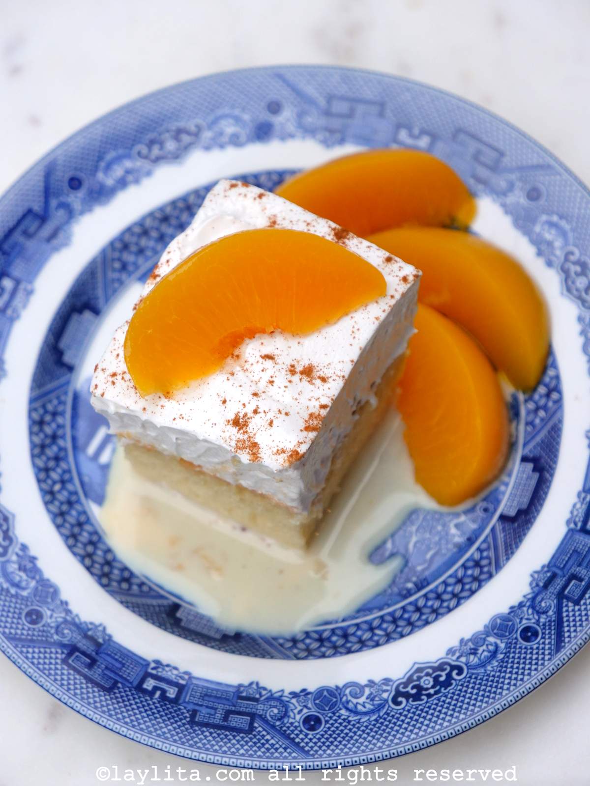 Slice of tres leches cake on a blue and white plate with peaches on top.