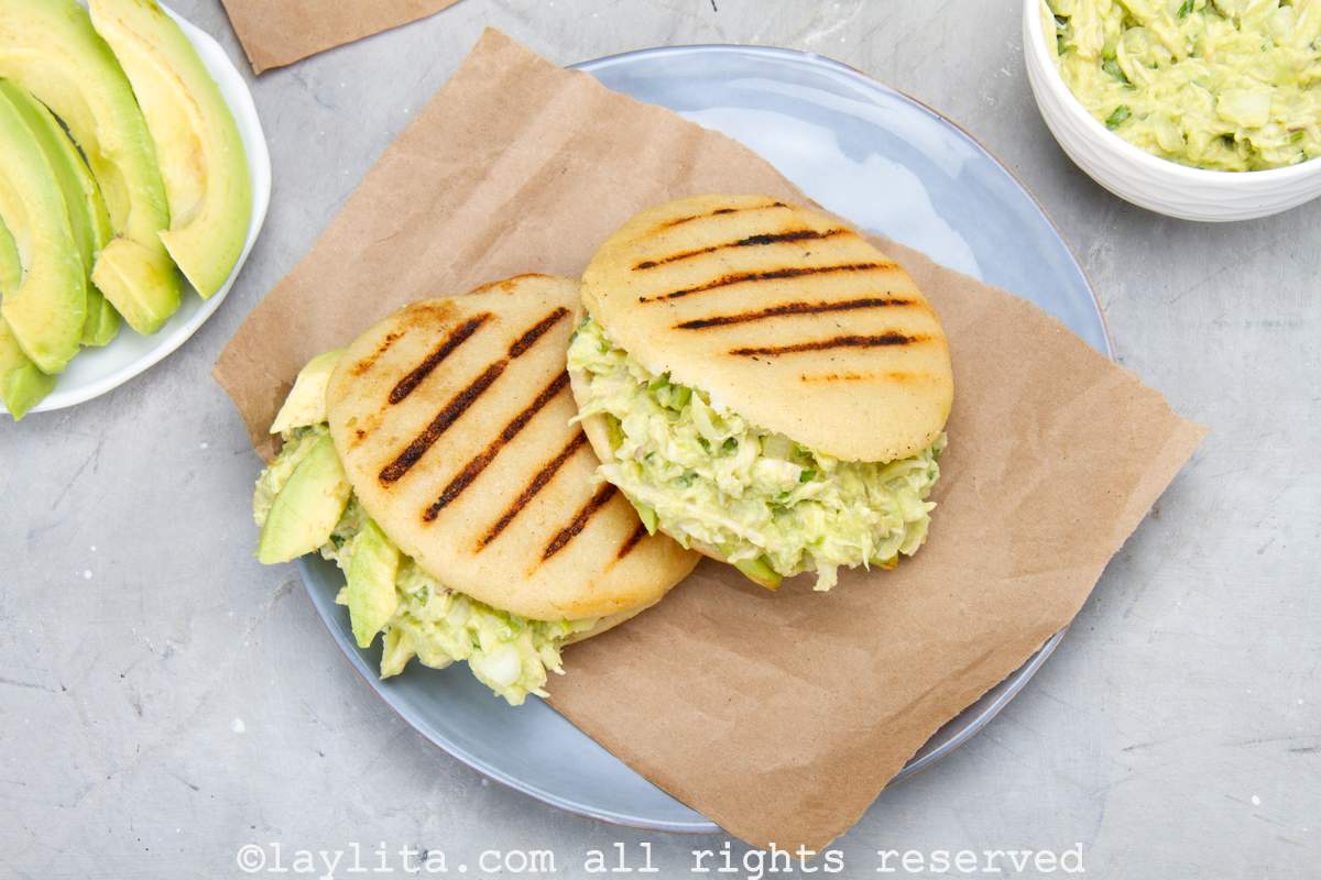Overhead shot of two reina pepiada arepas served on a plate with a plate of avocado and bowl of chicken avocado salad on the side.