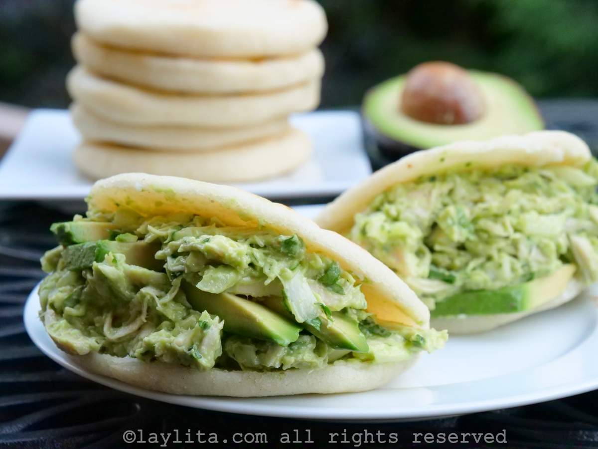 Outdoor photo of two Venezuelan style arepas filled with chicken avocado salad on a white plate, with a stack of arepas and an avocado half in the background