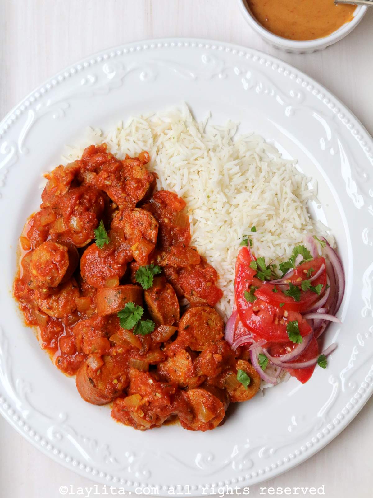A white plate with rougail sausage stew served with rice, tomato salad and peanut sauce.