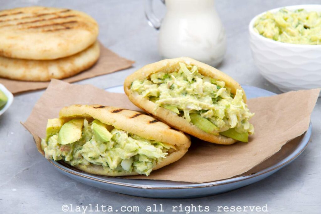 Two stuffed arepas served on a plate and stacked slightly on top of each other. A plate with arepas and mashed avocado in the background.