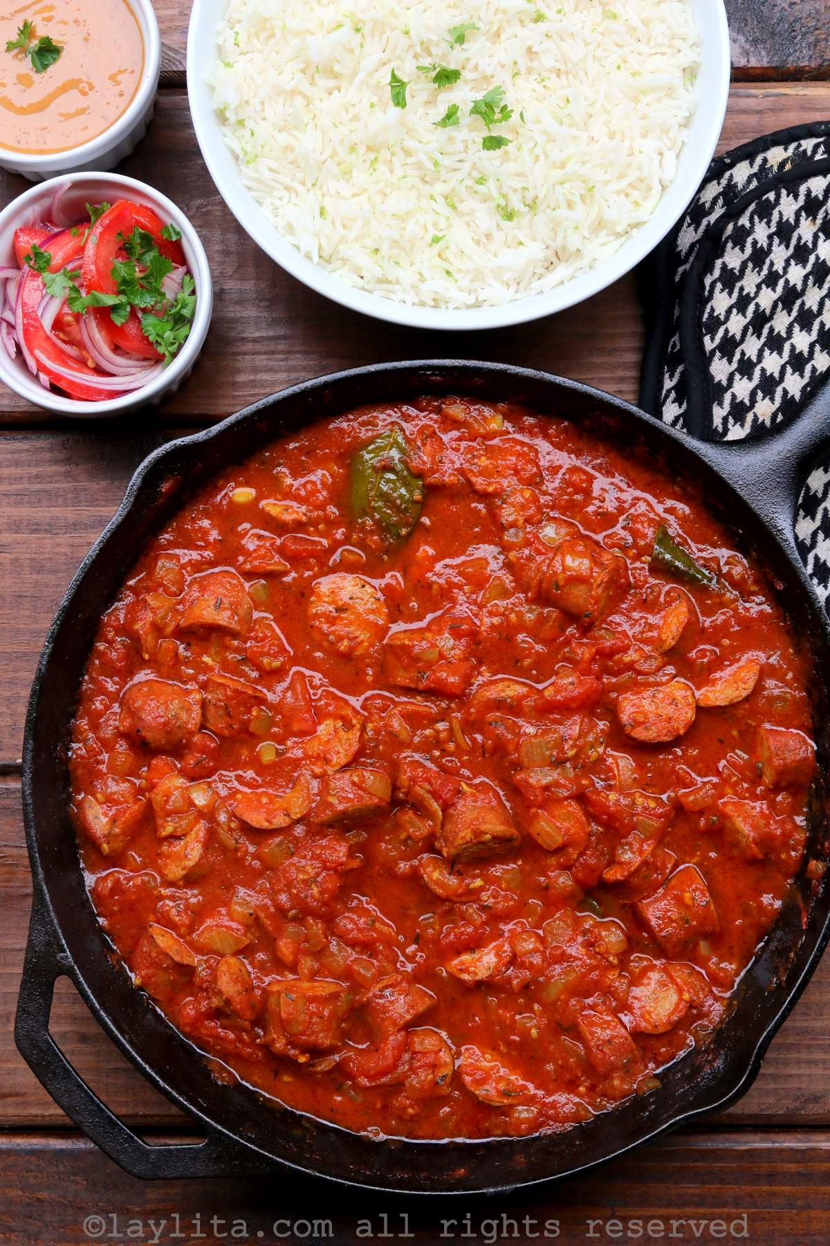 Rougail stew in a skillet on a wooden background with sides of rice, salad and peanut sauce on the side.