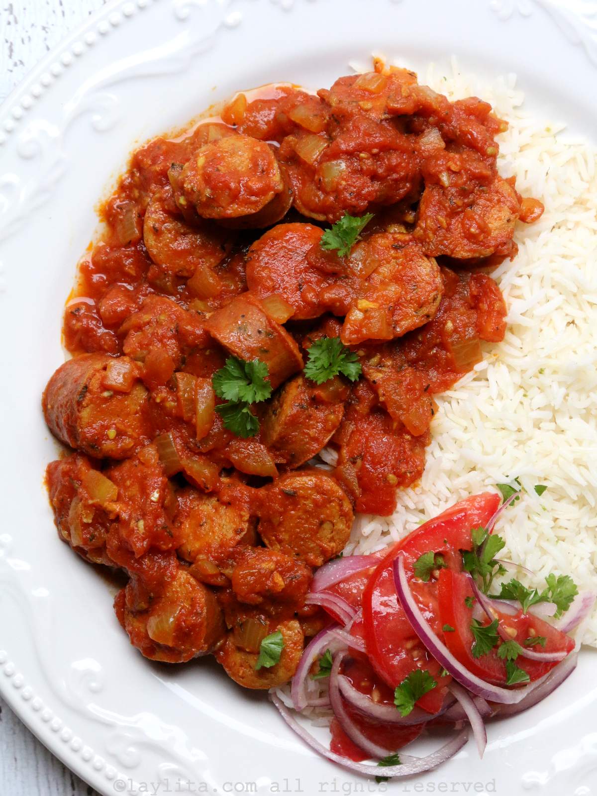 A white plate with rougaille saucisse sausage stew served with rice and tomato salad.