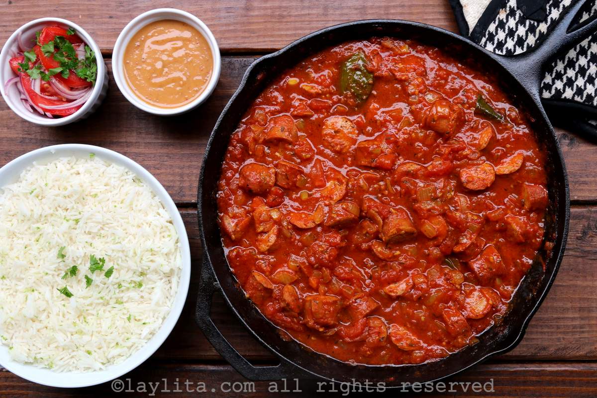 Rougail in a skillet with a bowl of rice, onions and peanut sauce next to it.