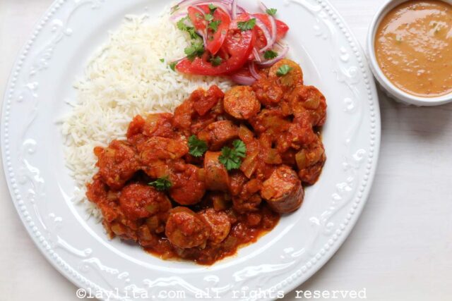 Rougail sausage stew served with rice in a white bowl with a side of tomatoes salad and peanut sauce.