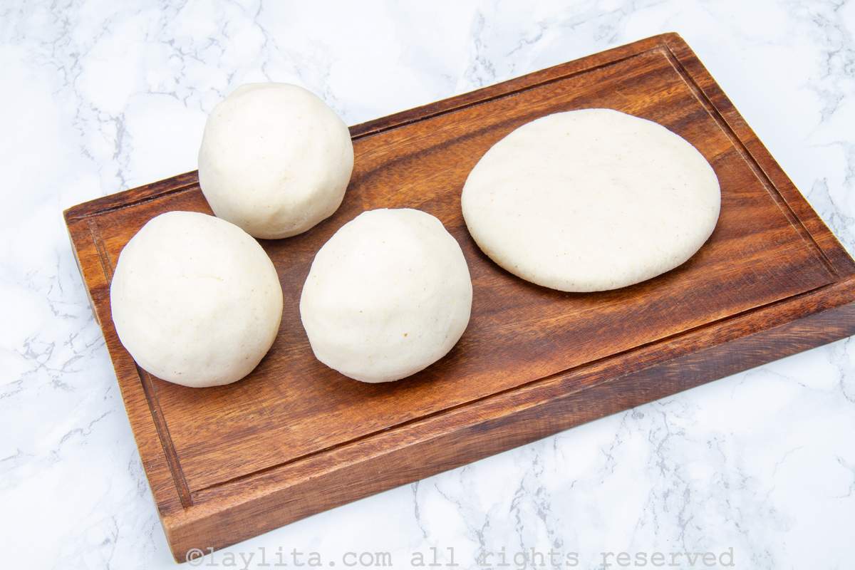 Three balls of arepa dough next to one that is shaped into a disc on a wooden cutting board.