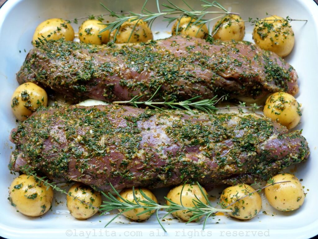Marinated pork tenderloins on a baking sheet with potatoes, onions and sprigs of herbs.