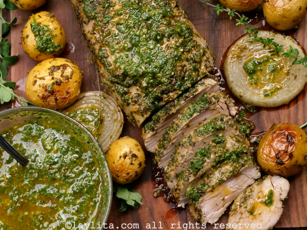 Pork tenderloin on a wooden cutting board with some slices off the end and more chimichurri on the side.