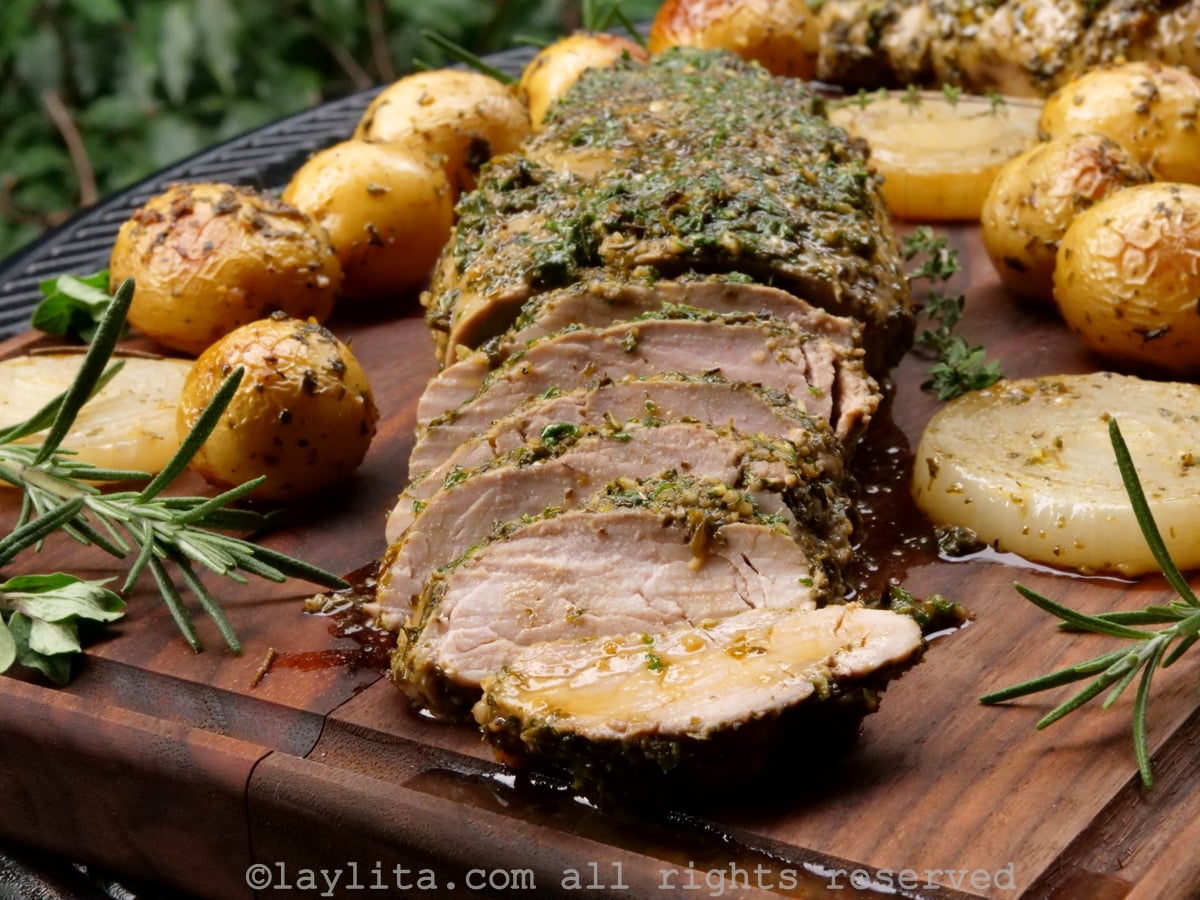 Baked pork tenderloin on a wooden background with some slices off the end, surrounded by roasted potatoes and onions.