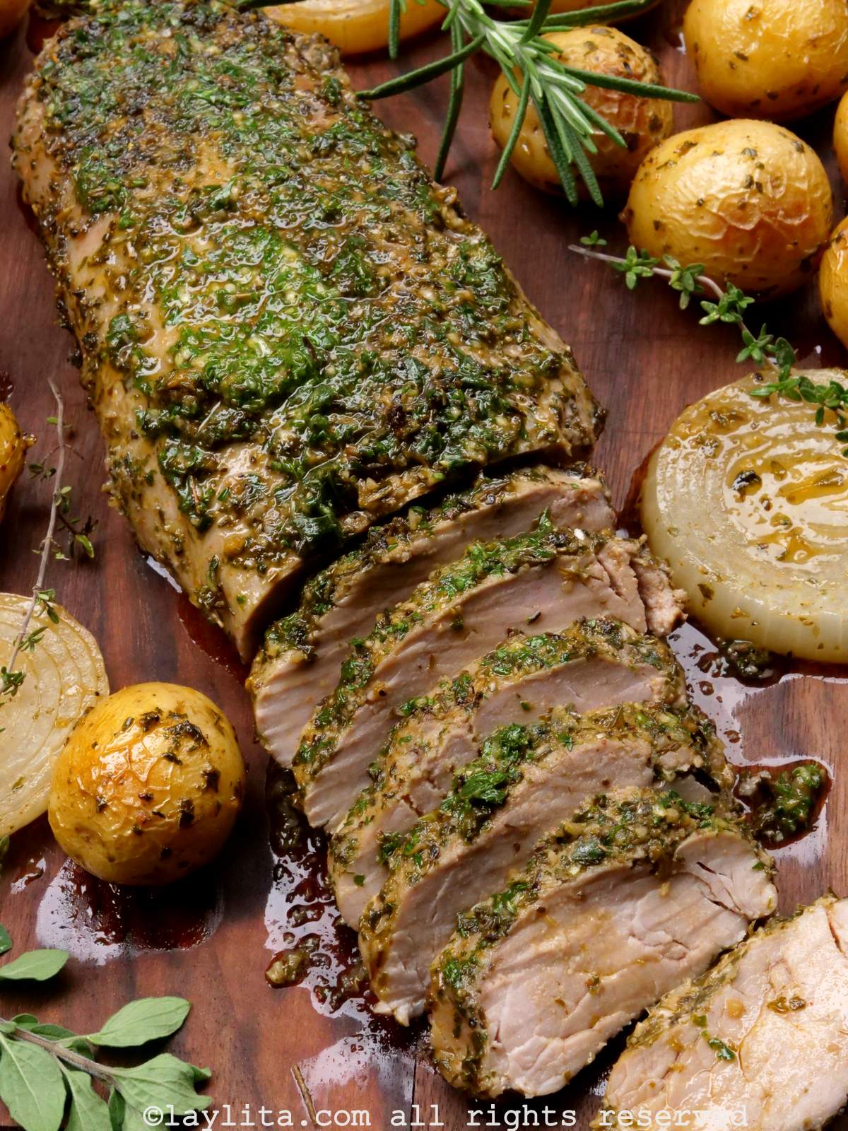 Overhead shot of a roasted pork tenderloin cut into slices and topped with chimichurri, surrounded by roasted potatoes, onions, and fresh herbs