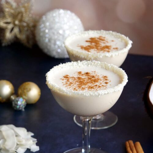 Two coupe glasses filled with Coquito eggnog and Christmas decorations in the background