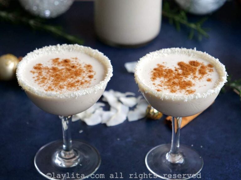 Two coupe glasses filled with Coquito eggnog and Christmas decorations and coconut slivers in the background