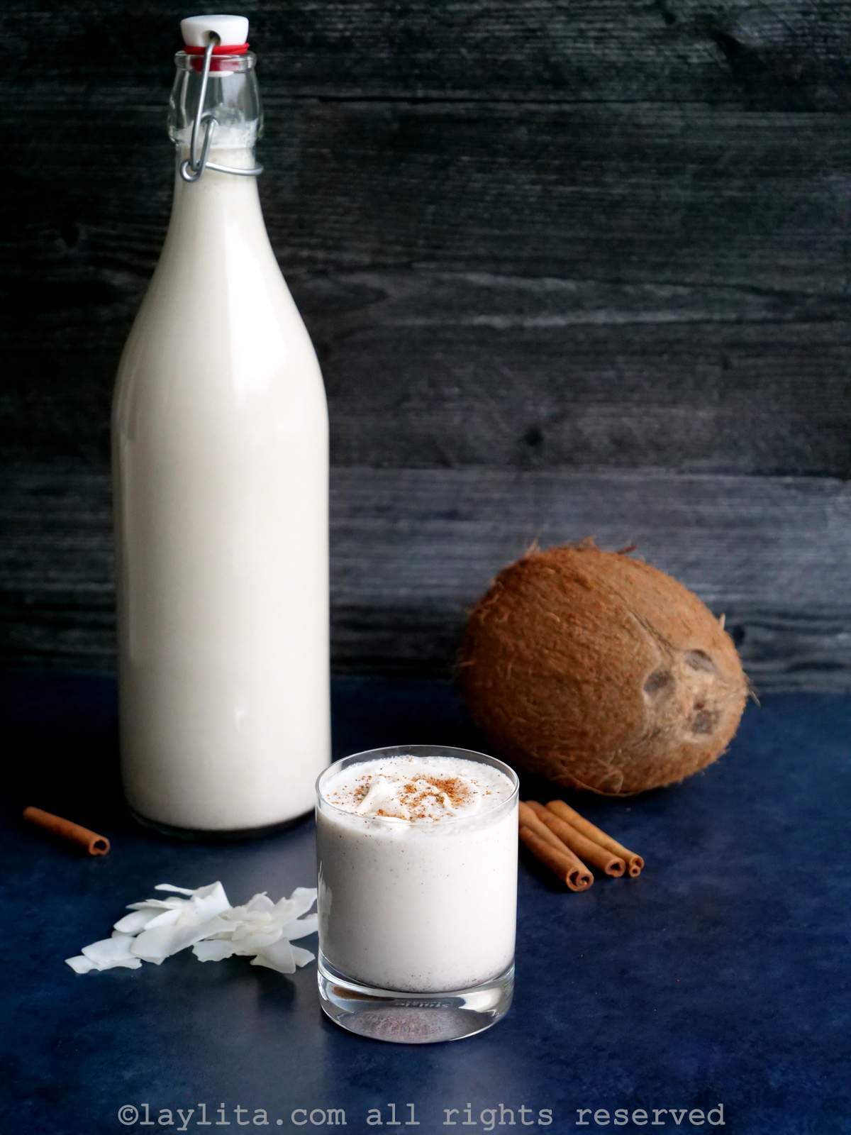 A glass of Coquito on a blue counter, with a bottle of Coquito and a coconut in the background