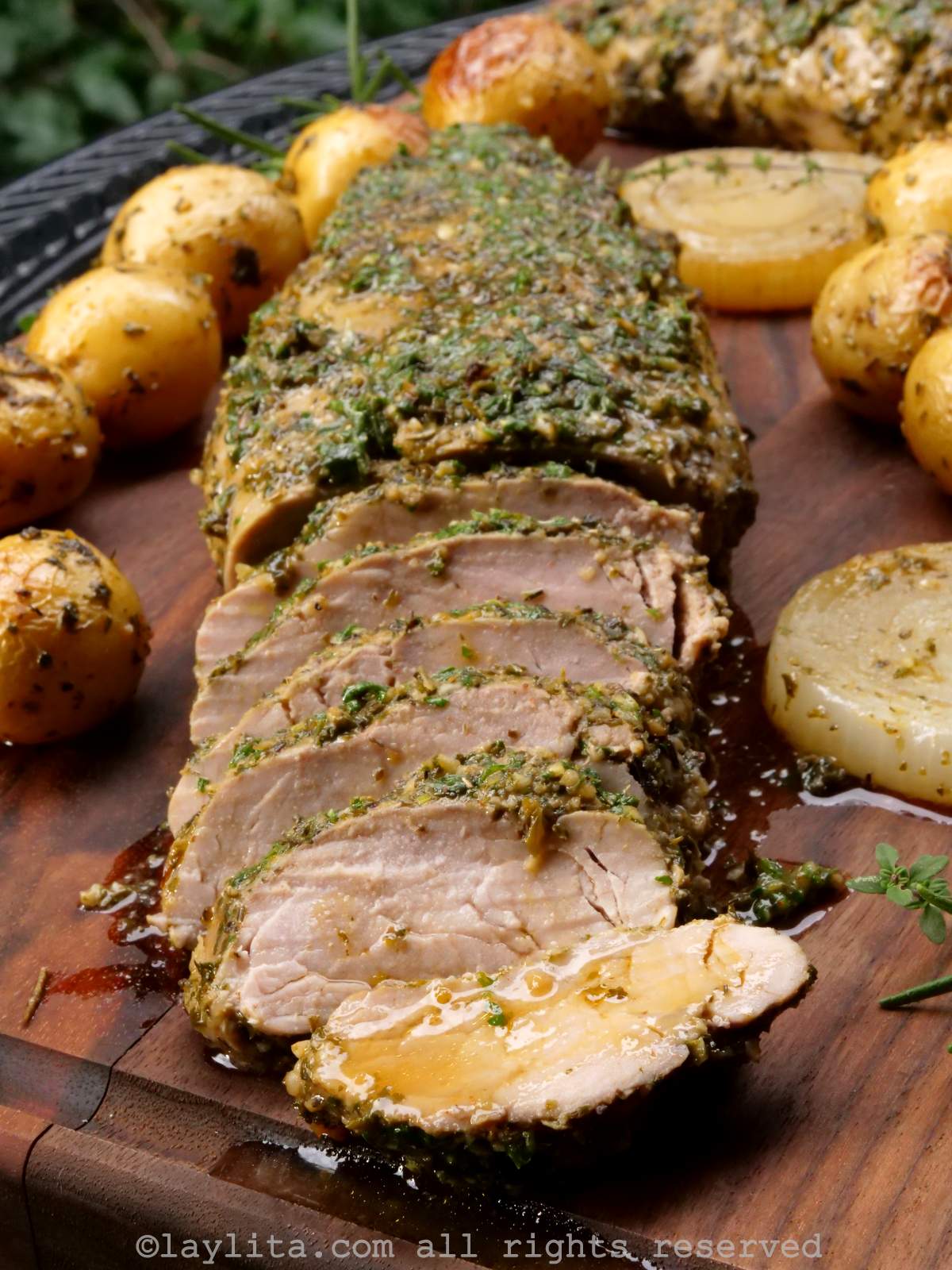 Roasted chimichurri pork tenderloin on a wooden background with some slices off the end, surrounded by roasted potatoes and onions.