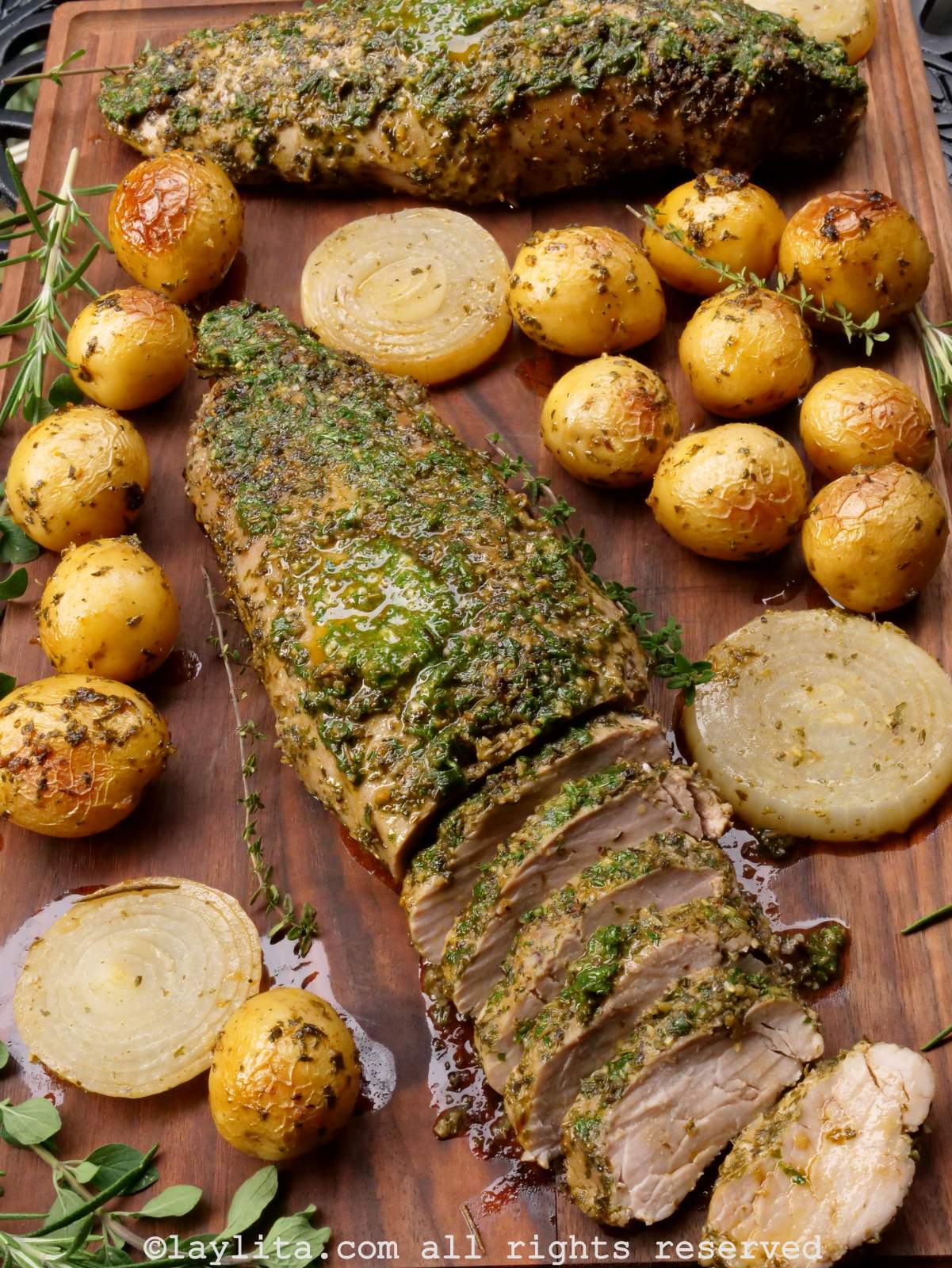 Overhead shot of two whole pork tenderloins on a wooden cutting board surrounded by roasted potatoes and onions.