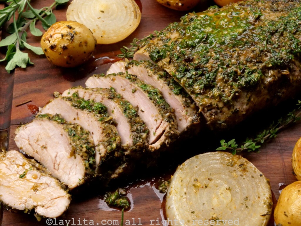 Side view of pork tenderloin on a wooden background with some slices off the end, surrounded by roasted potatoes and onions.