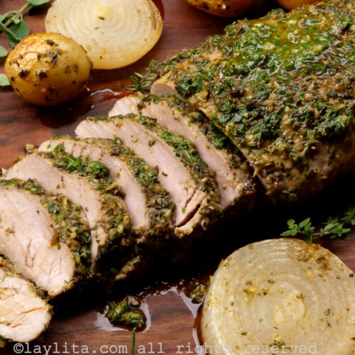 Side view of pork tenderloin on a wooden background with some slices off the end, surrounded by roasted potatoes and onions.