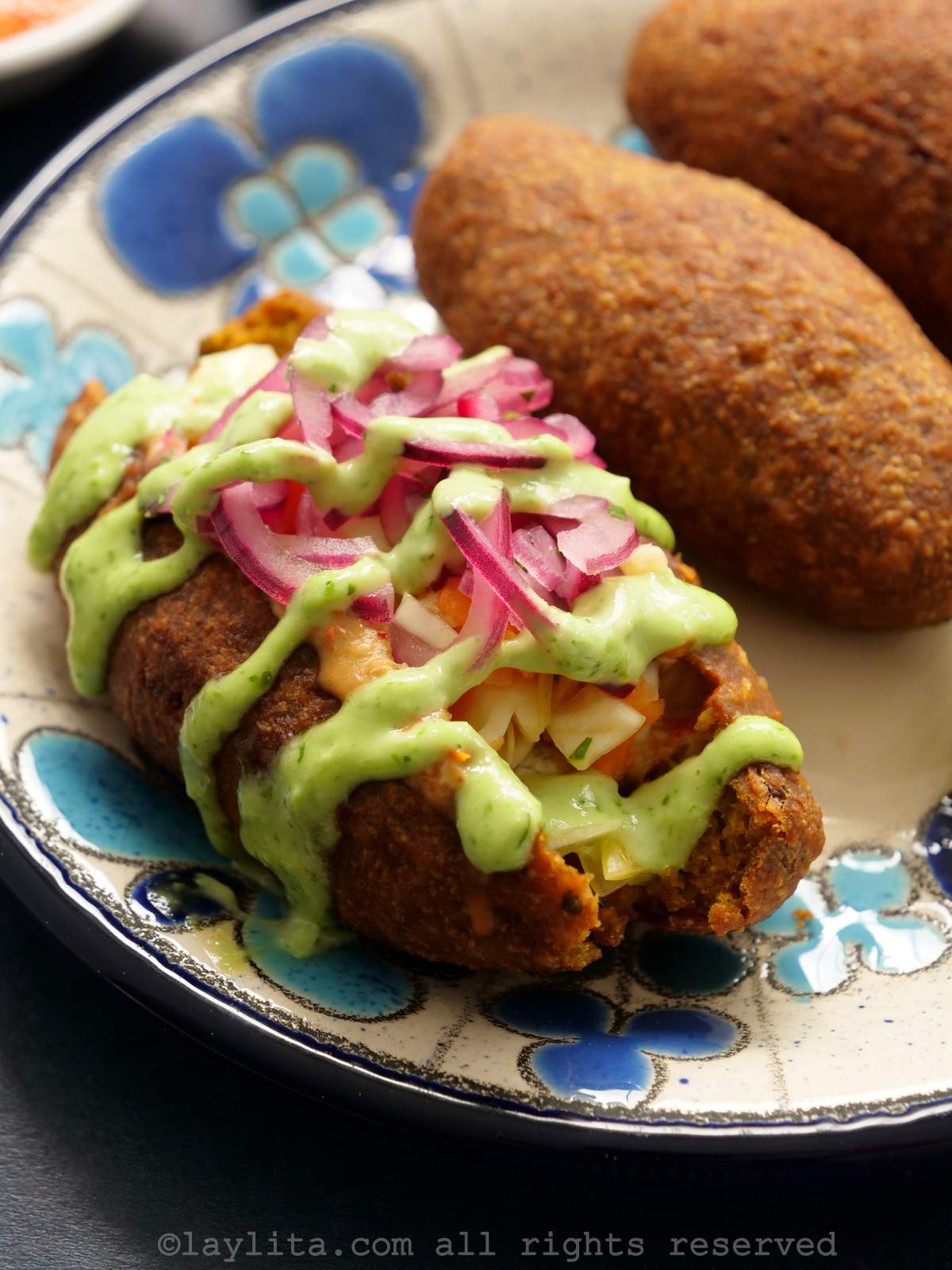 Close up shot of a corviche on a plate cut in half topped with onions and sauces with another corviche next to it.