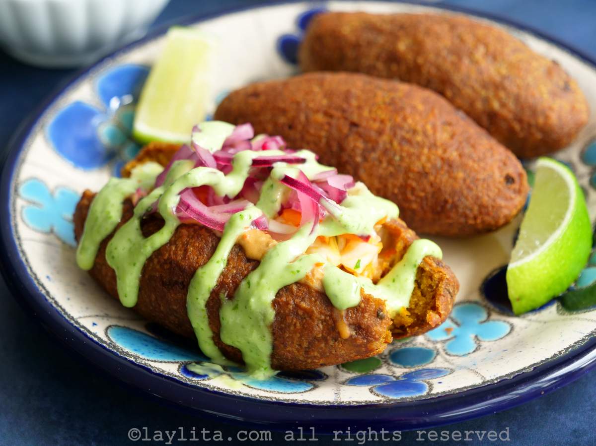 Close up shot of a corviche on a plate cut in half topped with onions and sauces with two more corviche croquettes next to it.