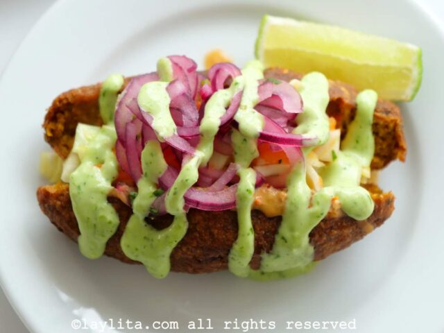 One corviche served on a plate with a drizzle of peanut sauce, green sauce, and pickled onions on top with a lime wedge on the side.
