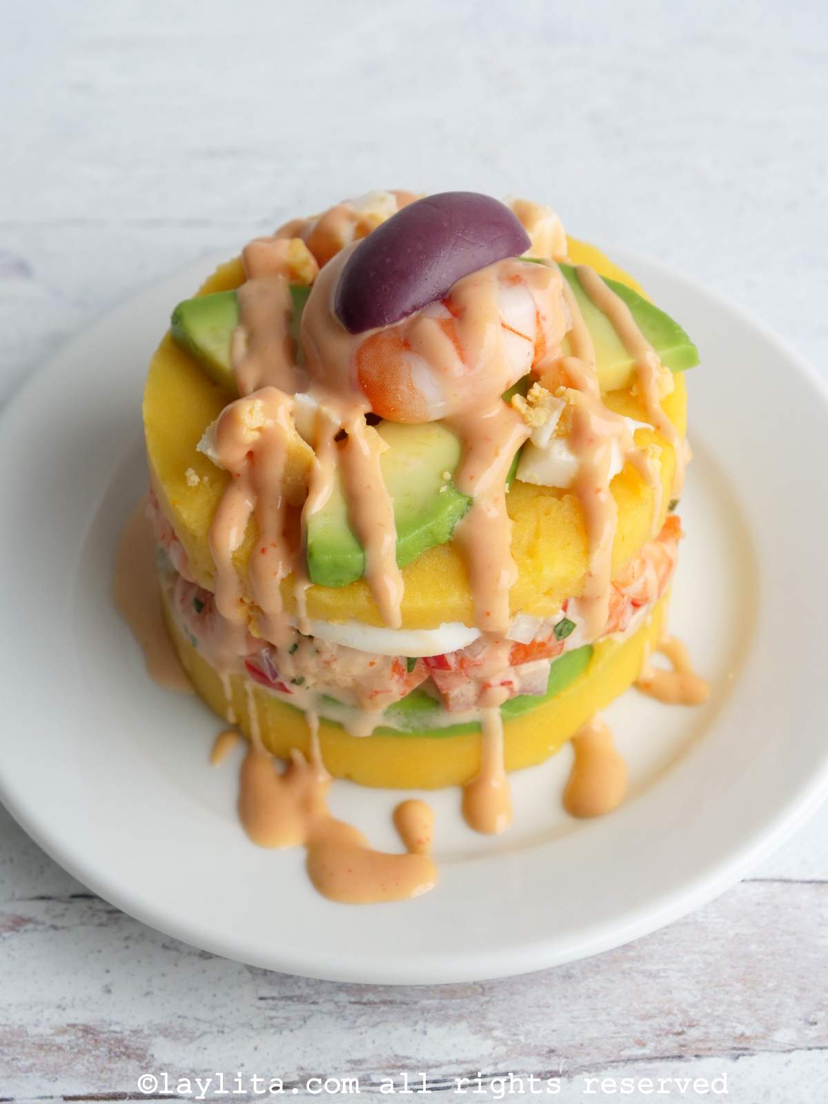 Overhead shot of a Peruvian causa de camarones topped with avocado, shrimp, purple olive, and a drizzle of pink sauce on a white plate