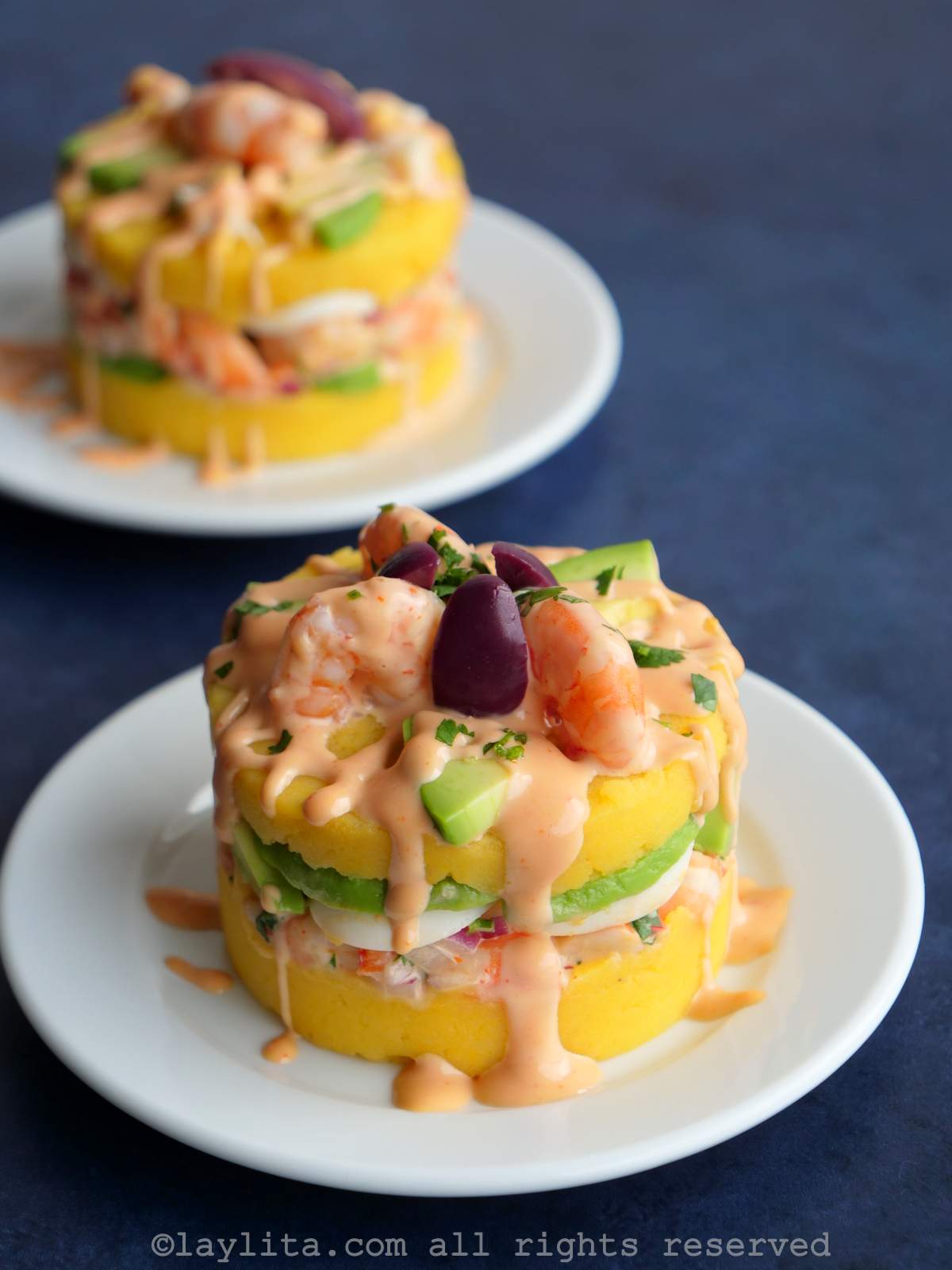 Overhead shot of a Peruvian causa de camarones topped with avocado, shrimp, purple olive, and a drizzle of pink sauce on a white plate with a blue background