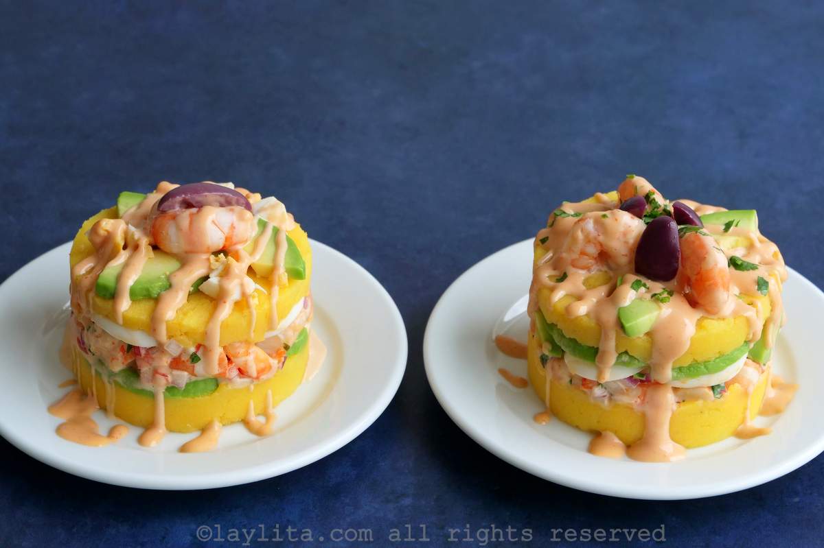 Two Peruvian causas de camarones topped with avocado, shrimp, purple olive, and a drizzle of pink sauce on white plates with a blue background