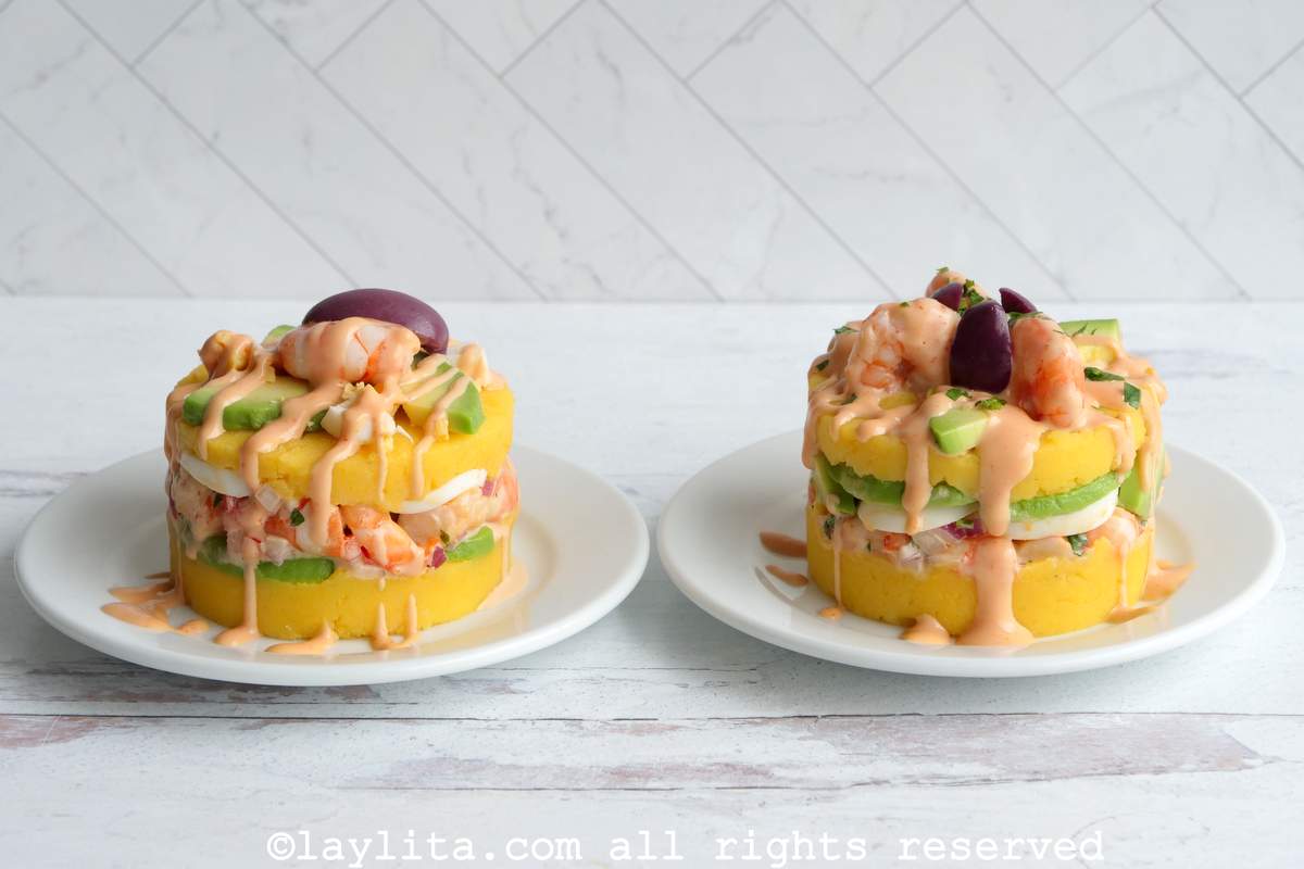 Causa de camarones served on a plate with shrimp, avocado and olives on top with a drizzle of pink sauce.