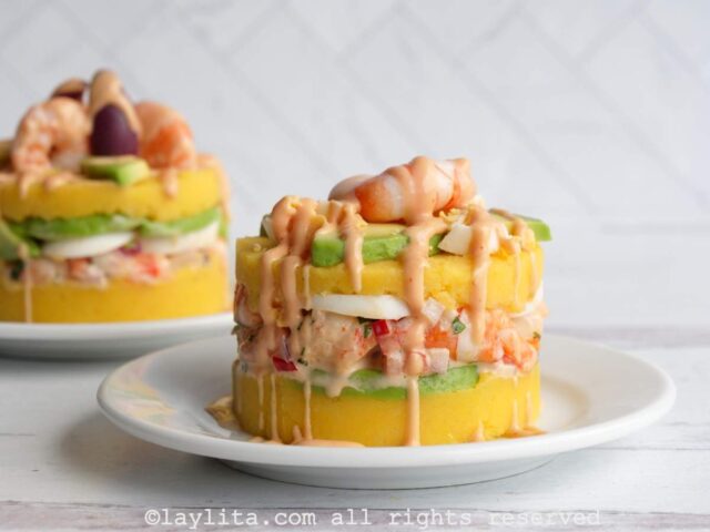 Causa de camarones served on a plate with shrimp, avocado and olives on top with a drizzle of pink sauce.