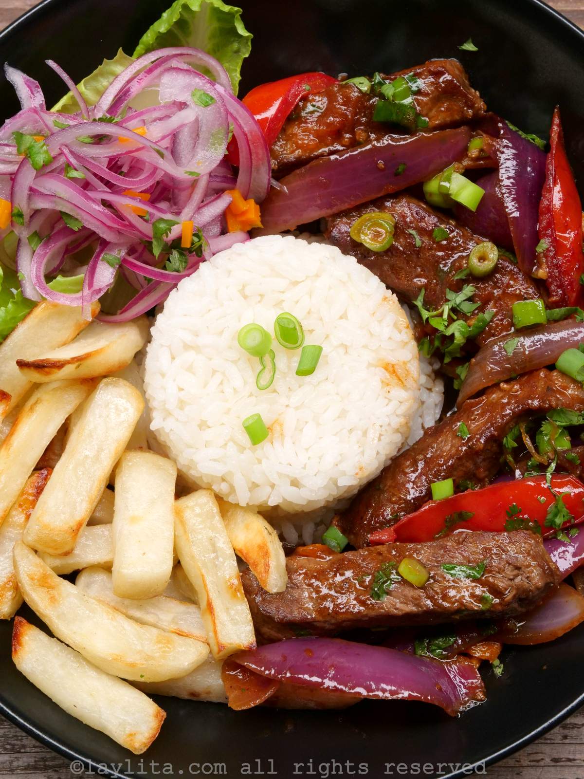 Overhead shot of the lomo saltado stir fry in black plate with rice, french fries and pickled red onions
