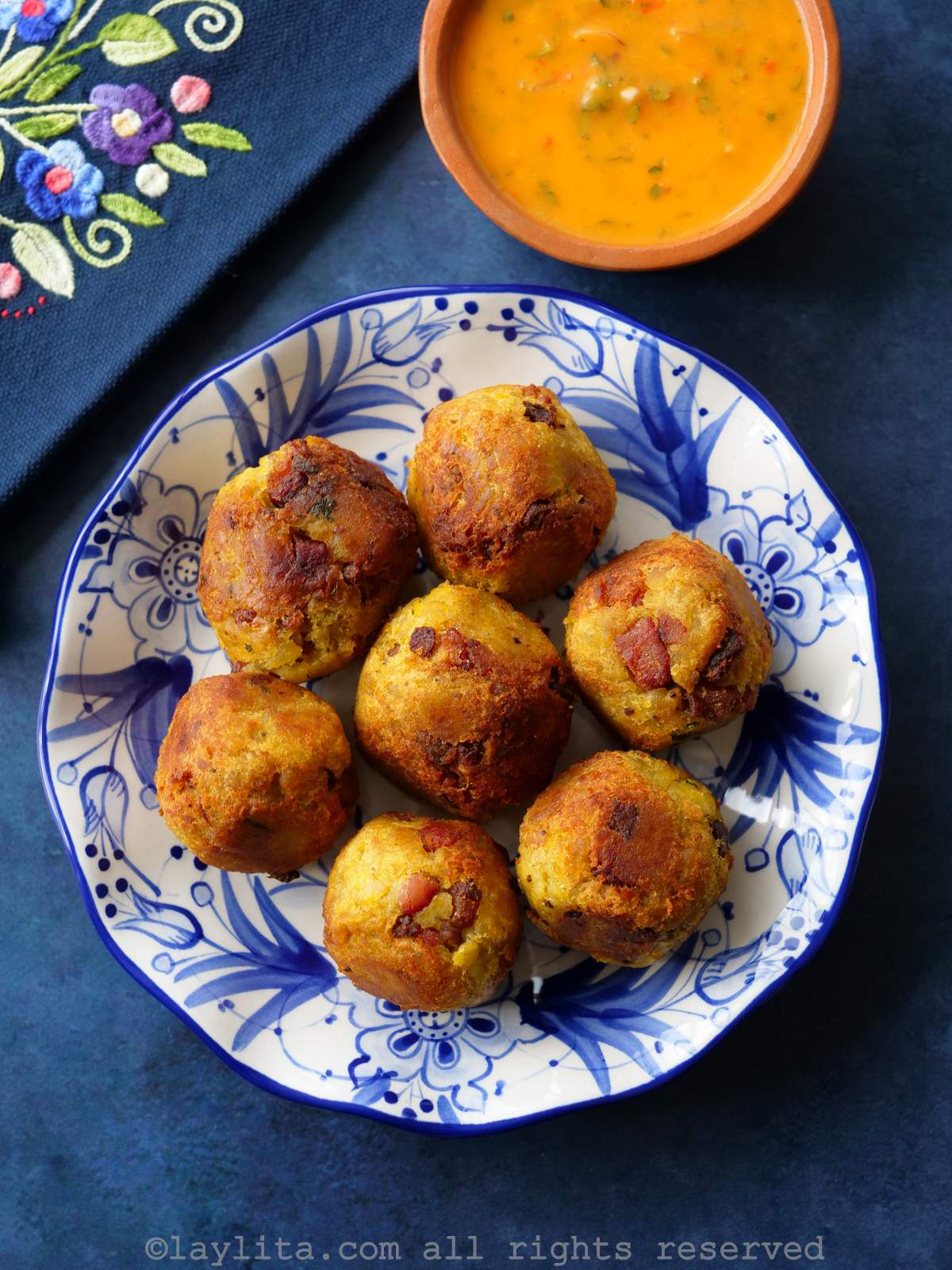 Bolon de verde or green plantain dumplings Bolones de verde or fried green plantain balls with crispy pork on a white and blue plate with a blue napkin and a bowl of hot sauce in the background
