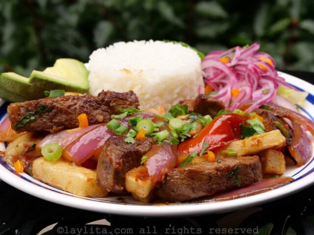 Lomo saltado served on a white plate on top of a bed of fries, with rice, onions and avocado.