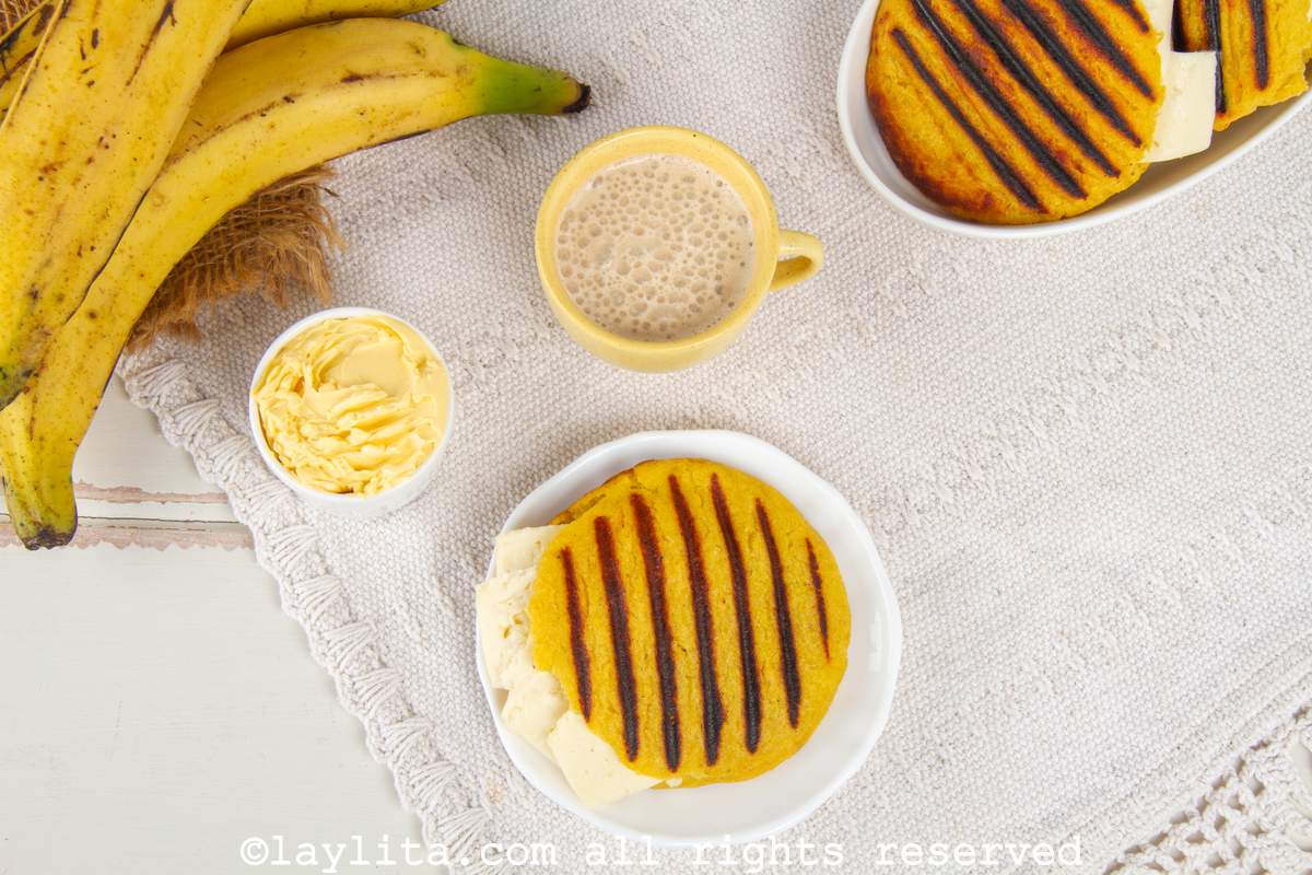 Overhead shot of an arepa de platano maduro on a white plate next to a cup of coffee, butter, plantains and more arepas.