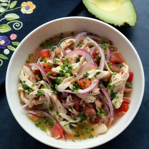 Ceviche de pollo in a white bowl with an avocado half and a colorful cloth napkin on the side