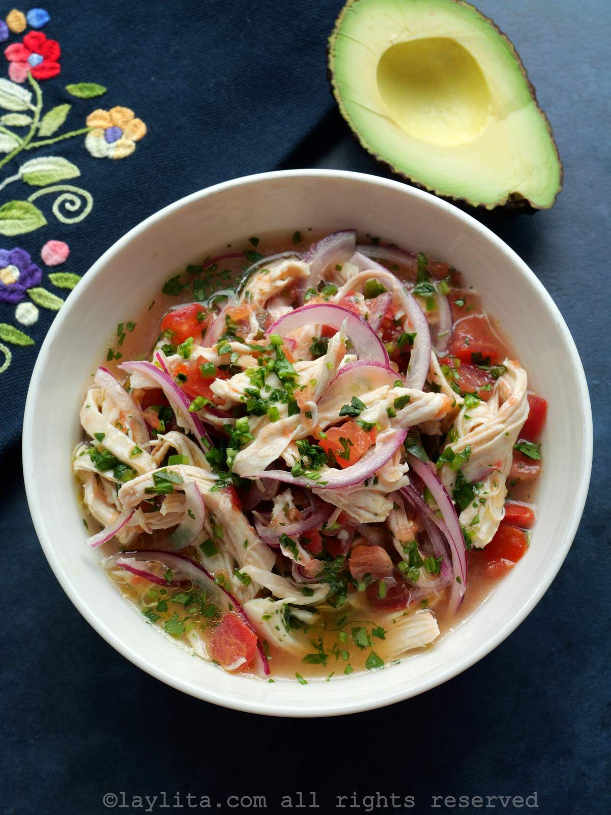Ceviche de pollo in a white bowl with an avocado half and a colorful cloth napkin on the side