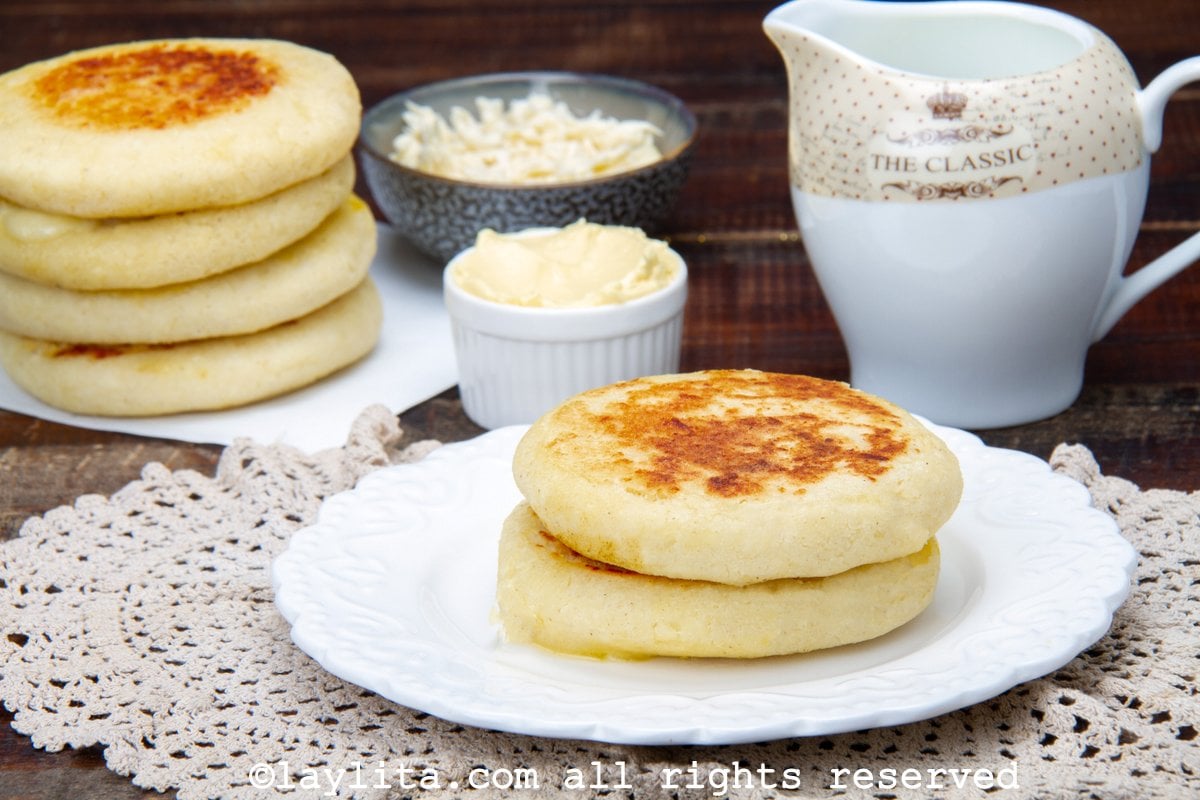 Two arepas on a plate and four arepas stacked on another plate, with a side of shredded cheese.