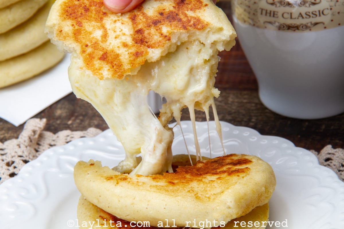 Two Colombian cheese arepas on a plate with one of the broken in half and a half pulling on it to show the cheese.