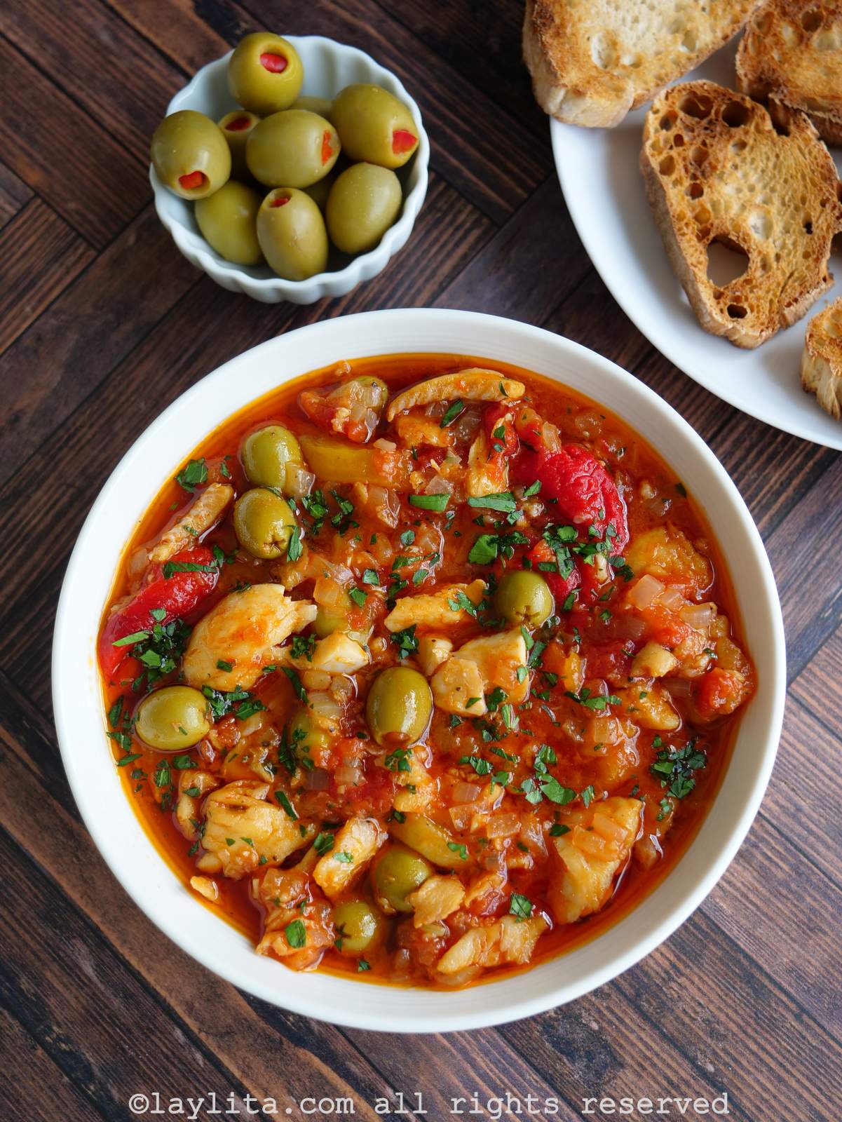 Bacalao a la vizcaina served in a white bowl next to a plate of toasted slices of bread, olives, and capers.