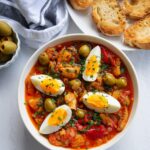 Bacalao a la vizcaina served in a white bowl next to a plate of toasted slices of bread, olives, and capers.