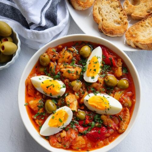 Bacalao a la vizcaina served in a white bowl next to a plate of toasted slices of bread, olives, and capers.