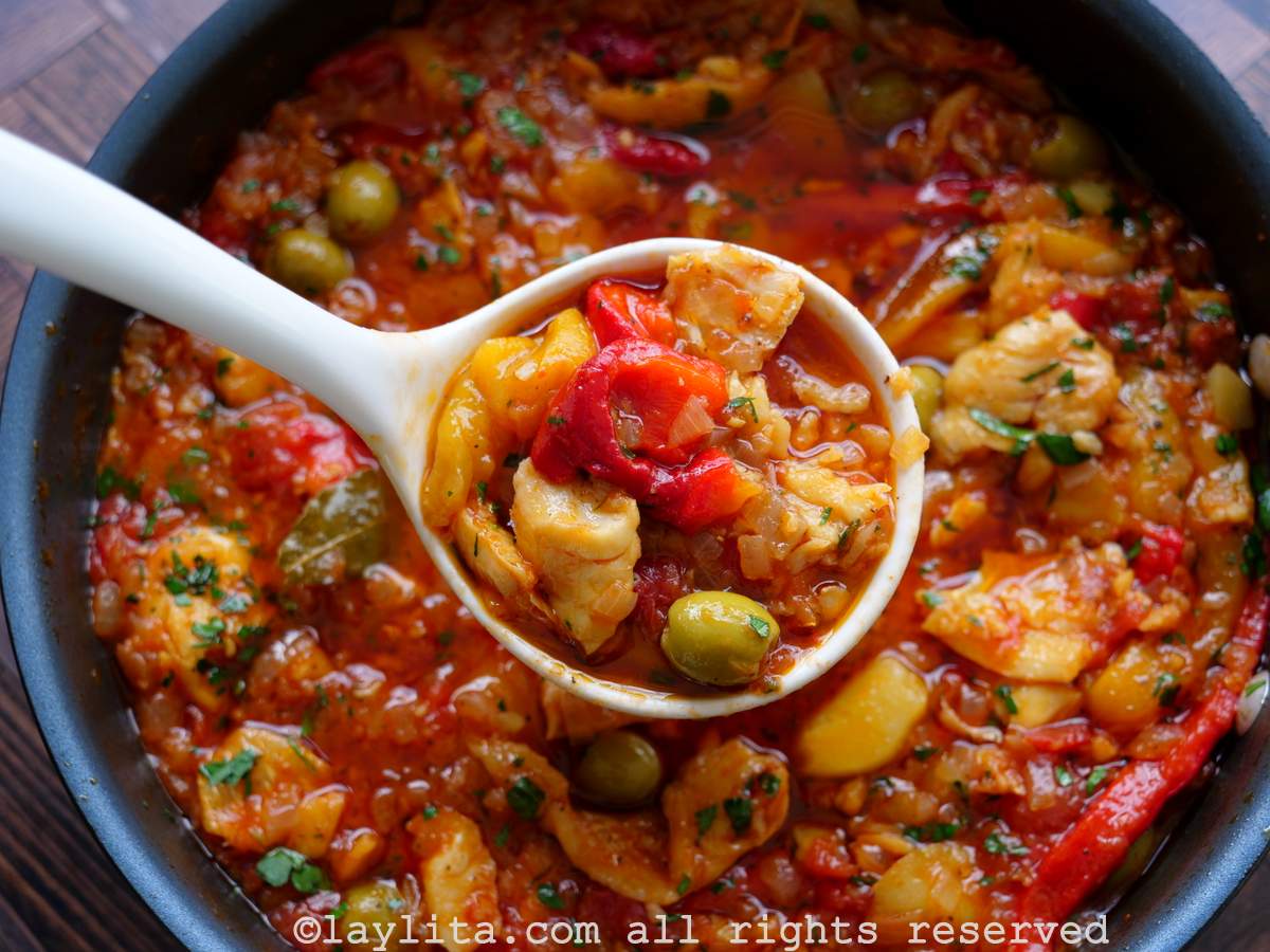 Close up of salted cod fish in a tomato and roasted pepper sauce with a ladle scooping up a portion from the pot.