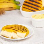 Ripe plantain arepa on a plate with sliced cheese stuffed in the middle and more plantains and arepas in the background.