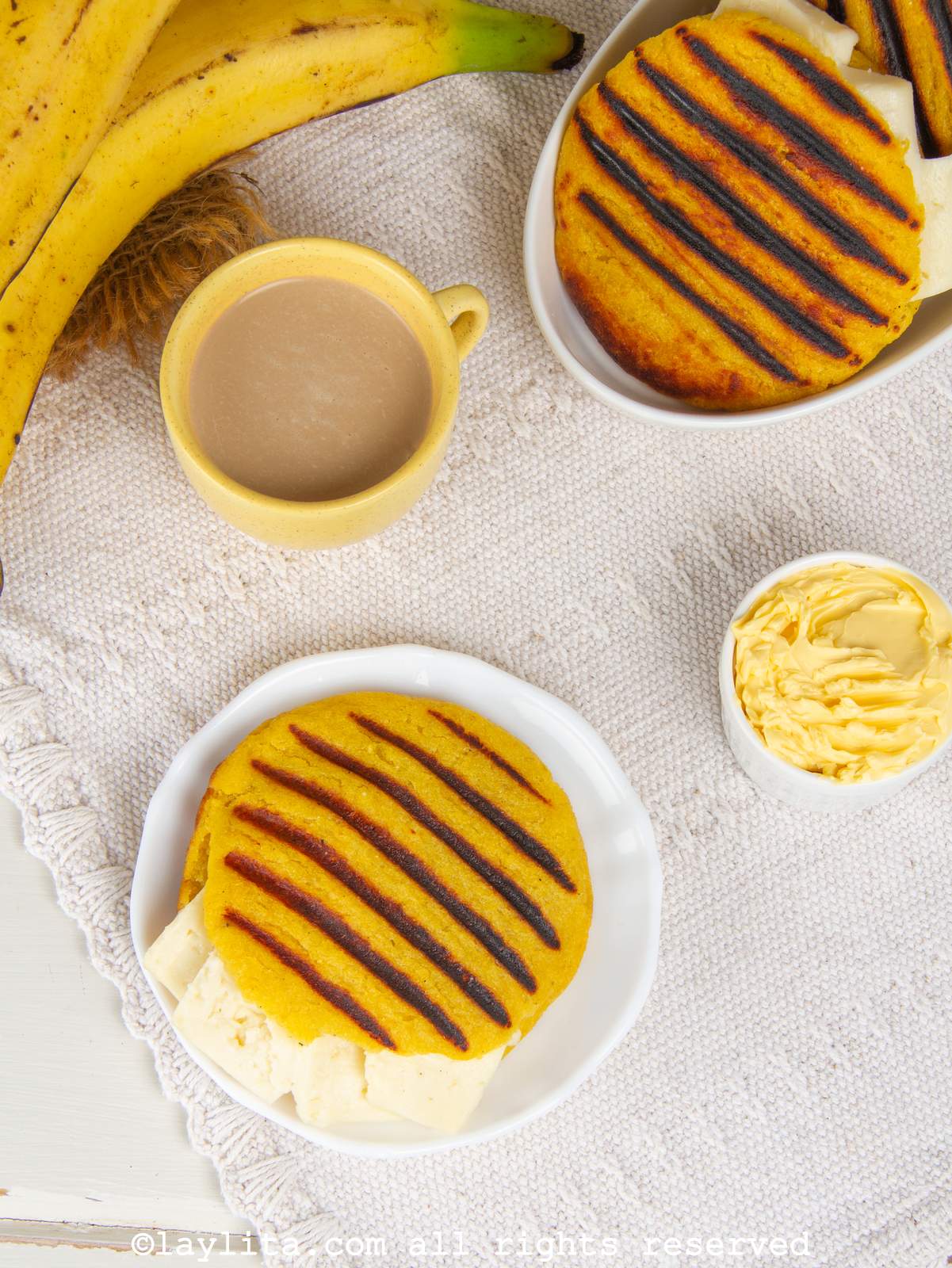 Overhead shot of an arepa de platano maduro on a white plate next to a cup of coffee, butter, plantains and more arepas.